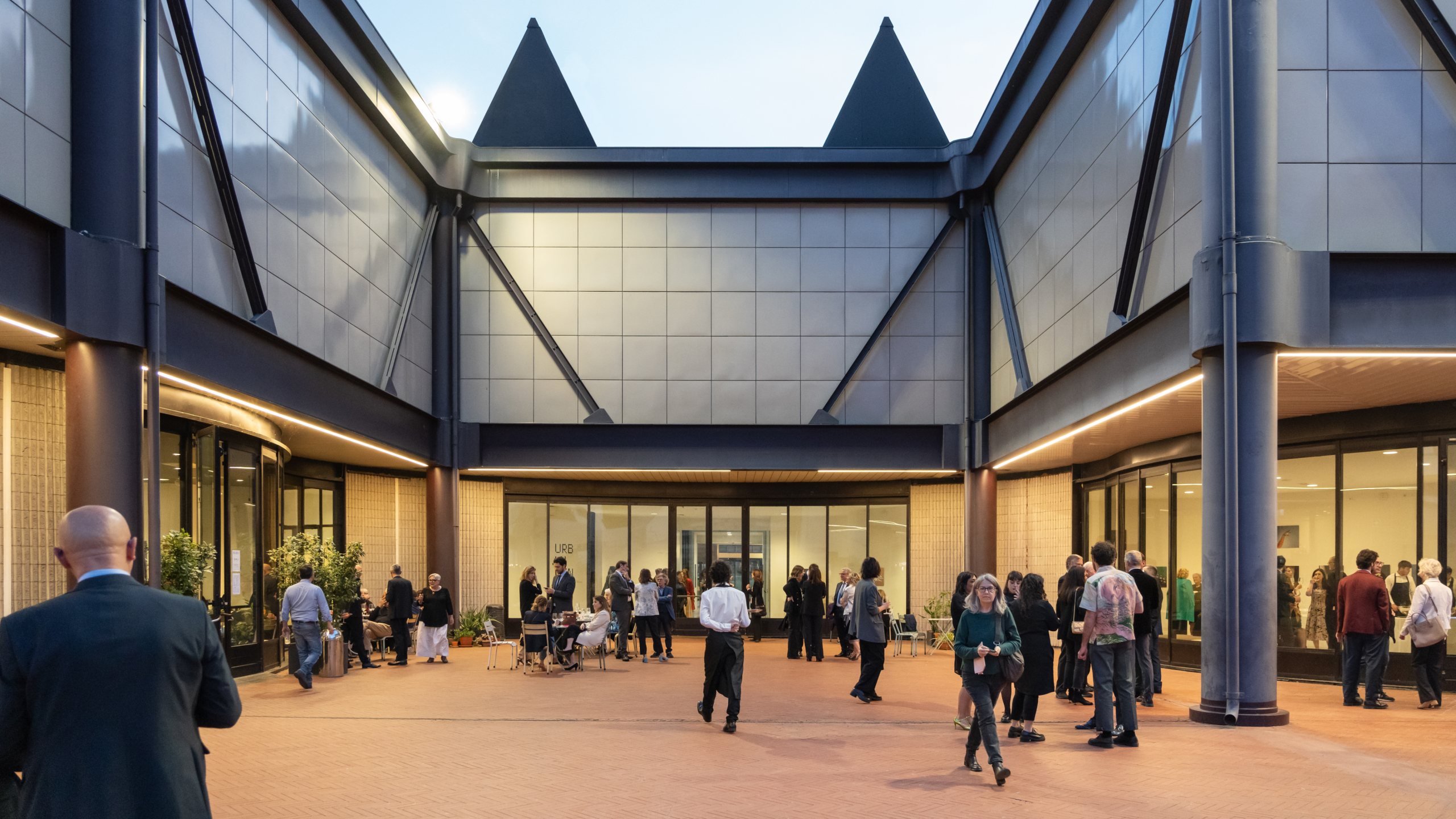Persone riunite nel cortile interno del Centro Pecci al crepuscolo, tra vetrate illuminate e architetture moderne, durante un evento serale.