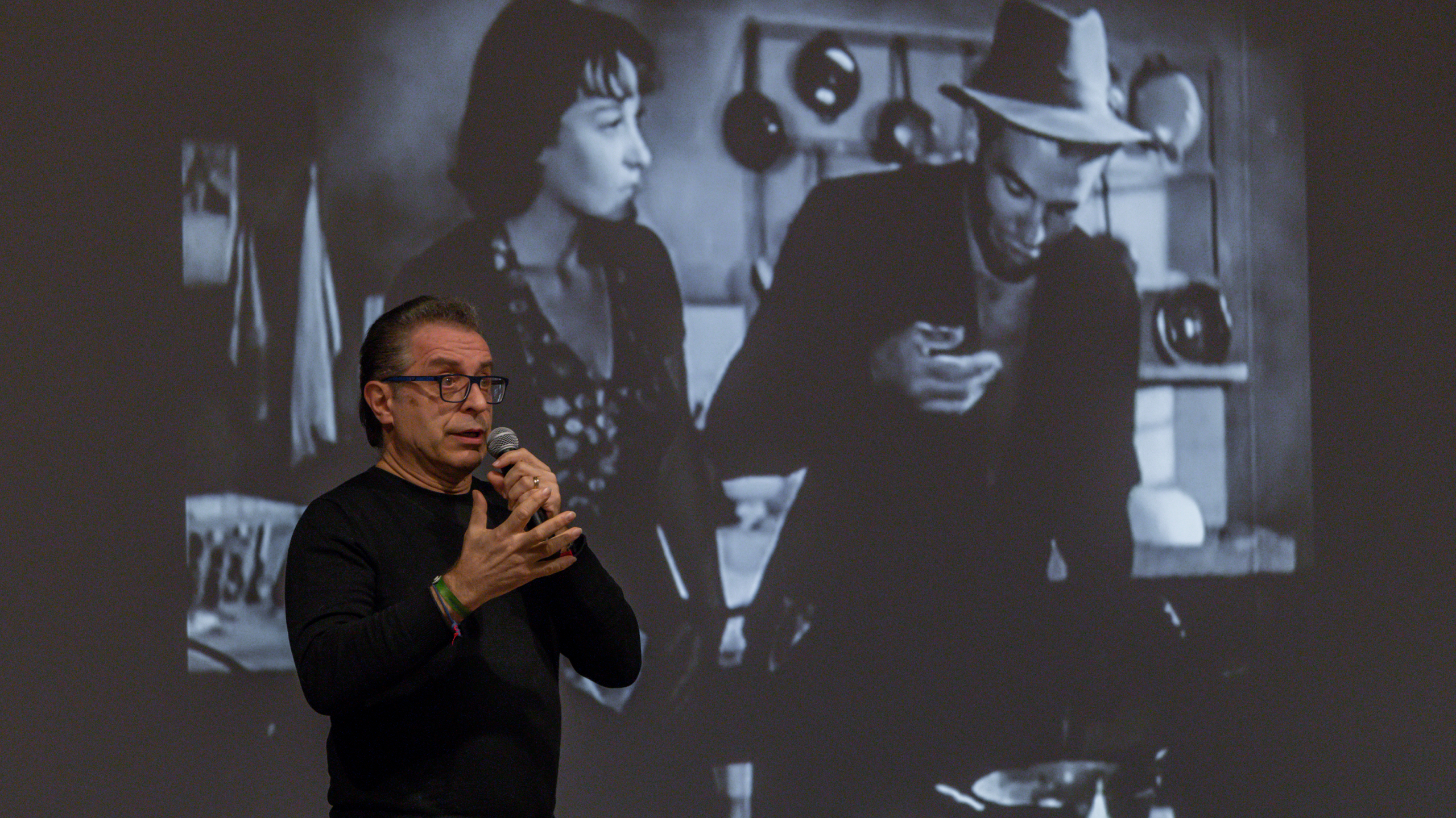 A speaker with a microphone talks in front of a screen projecting a black-and-white film scene during a cinema talk.