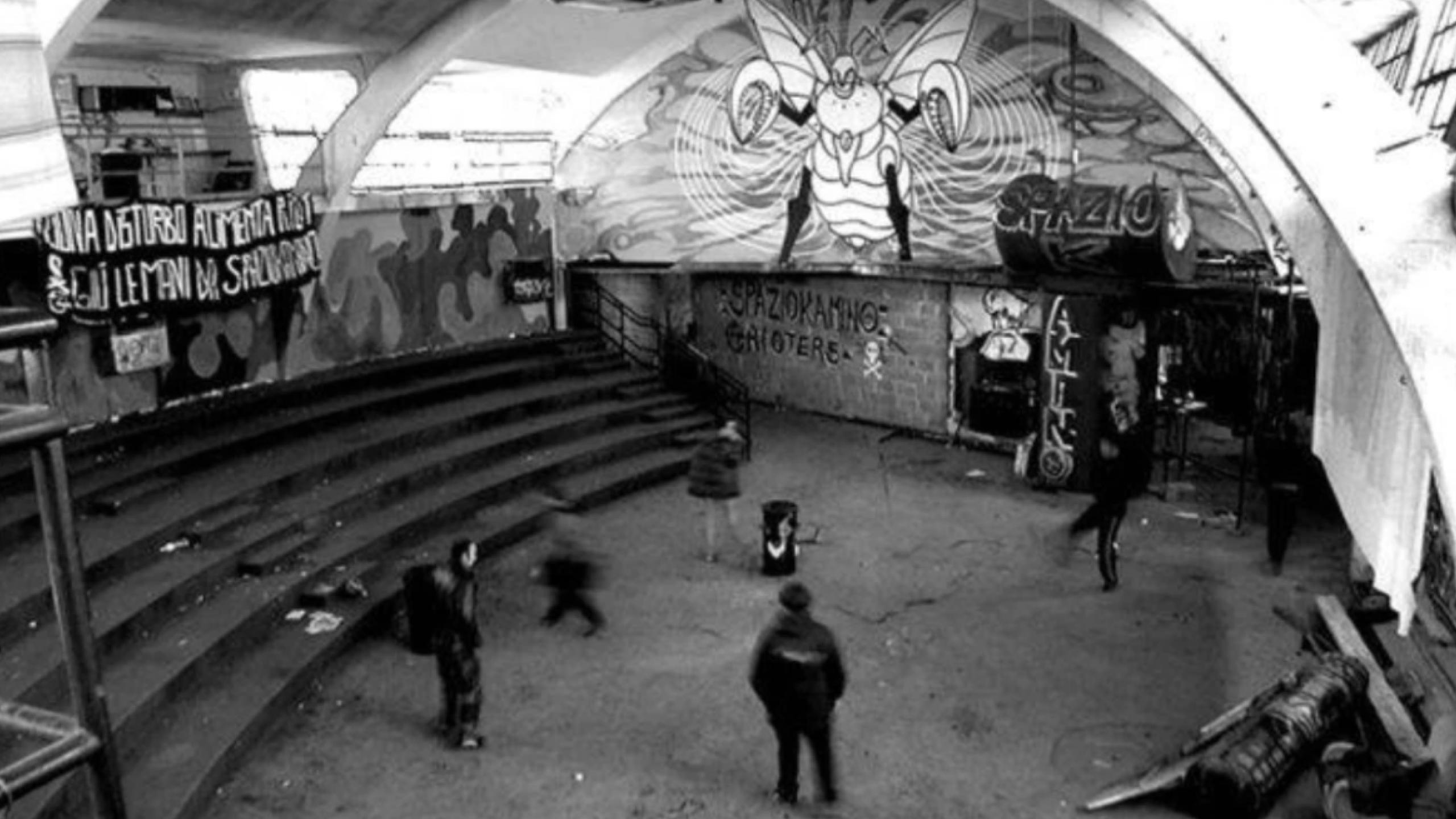 Vista in bianco e nero di uno spazio urbano coperto con gradinate laterali e muri ricoperti di graffiti e murales. Alcune persone si muovono nello spazio centrale, mentre sul fondo si vede un grande murale stilizzato e varie scritte sui muri.