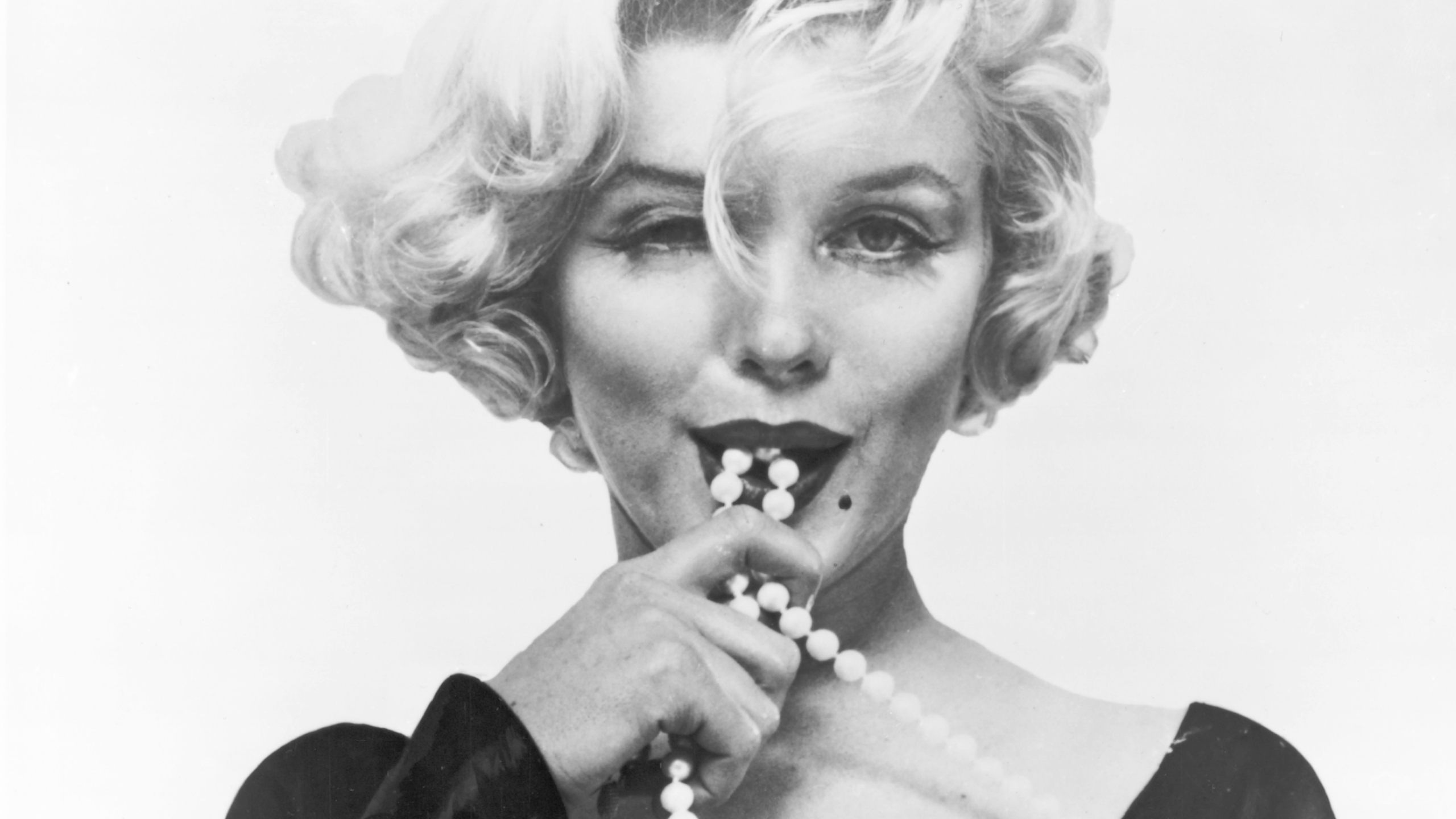 Black-and-white close-up portrait photograph of a person with short wavy hair, looking directly at the camera while holding a strand of pearls near the mouth.